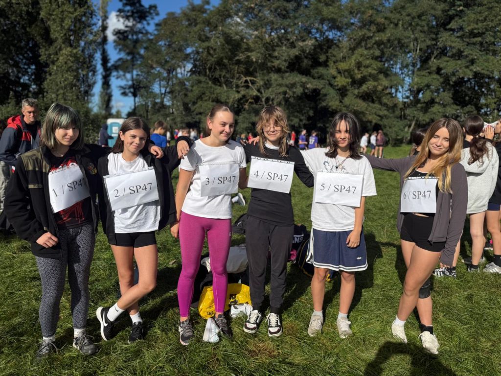 Sześć dziewcząt w sportowych strojach stoi na trawie podczas zawodów biegowych. Mają przy-pięte numery startowe z oznaczeniem SP47. Uśmiechają się i obejmują ramionami, tworząc zwartą grupę. W tle widać innych uczestników oraz drzewa w słoneczny dzień.