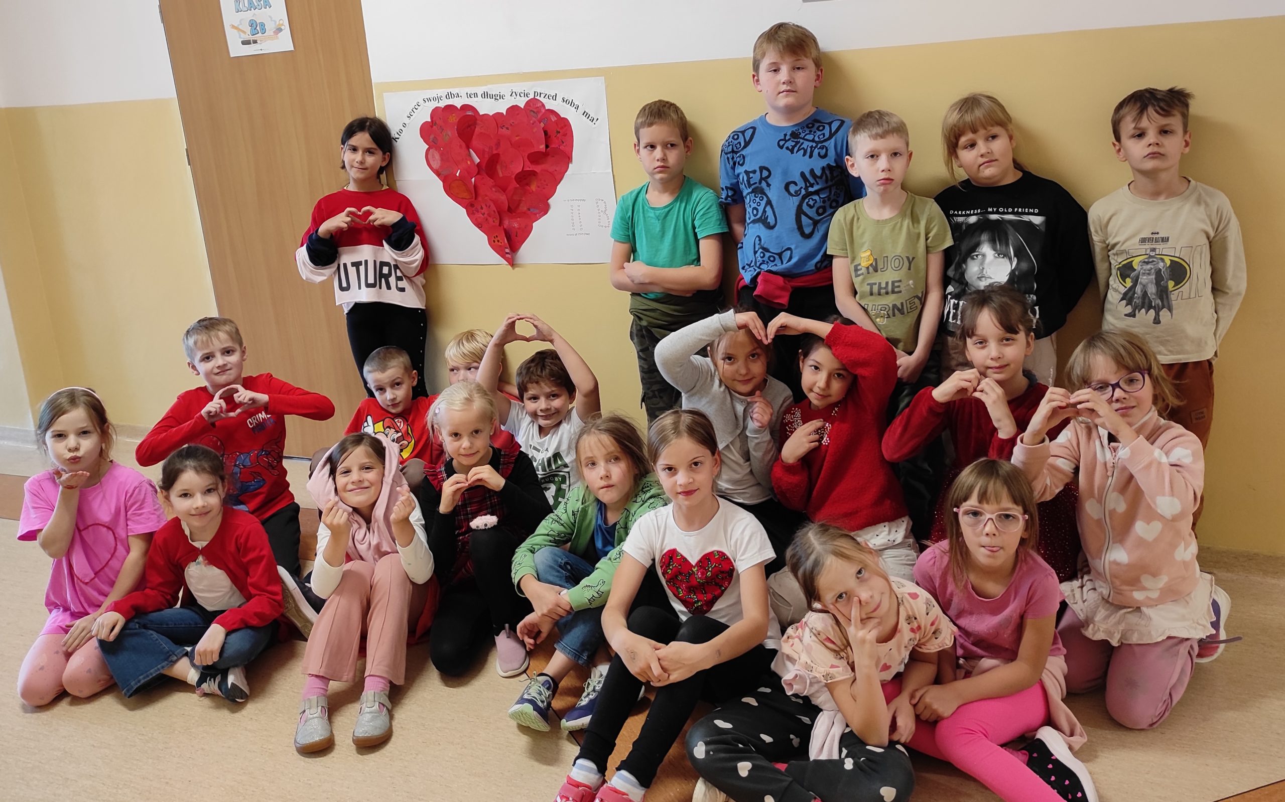 22 uczniów klasy 2b pozuje z dłońmi ułożonymi w kształt serca. Na ścianie plakat, do którego przyklejone są papierowe serca, ułożone w duże serce.