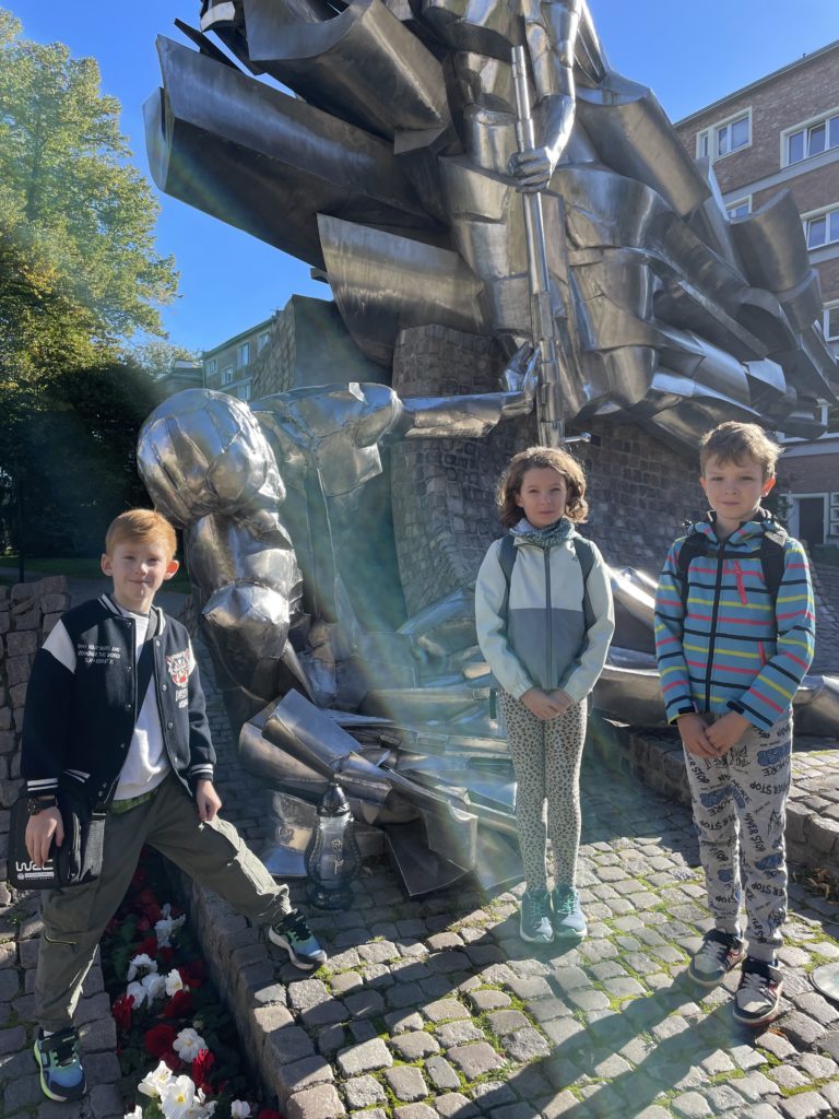 Dwóch chłopców i jedna dziewczynka stoją pod metalowym Pomnikiem Obrońców Poczty Polskiej. 