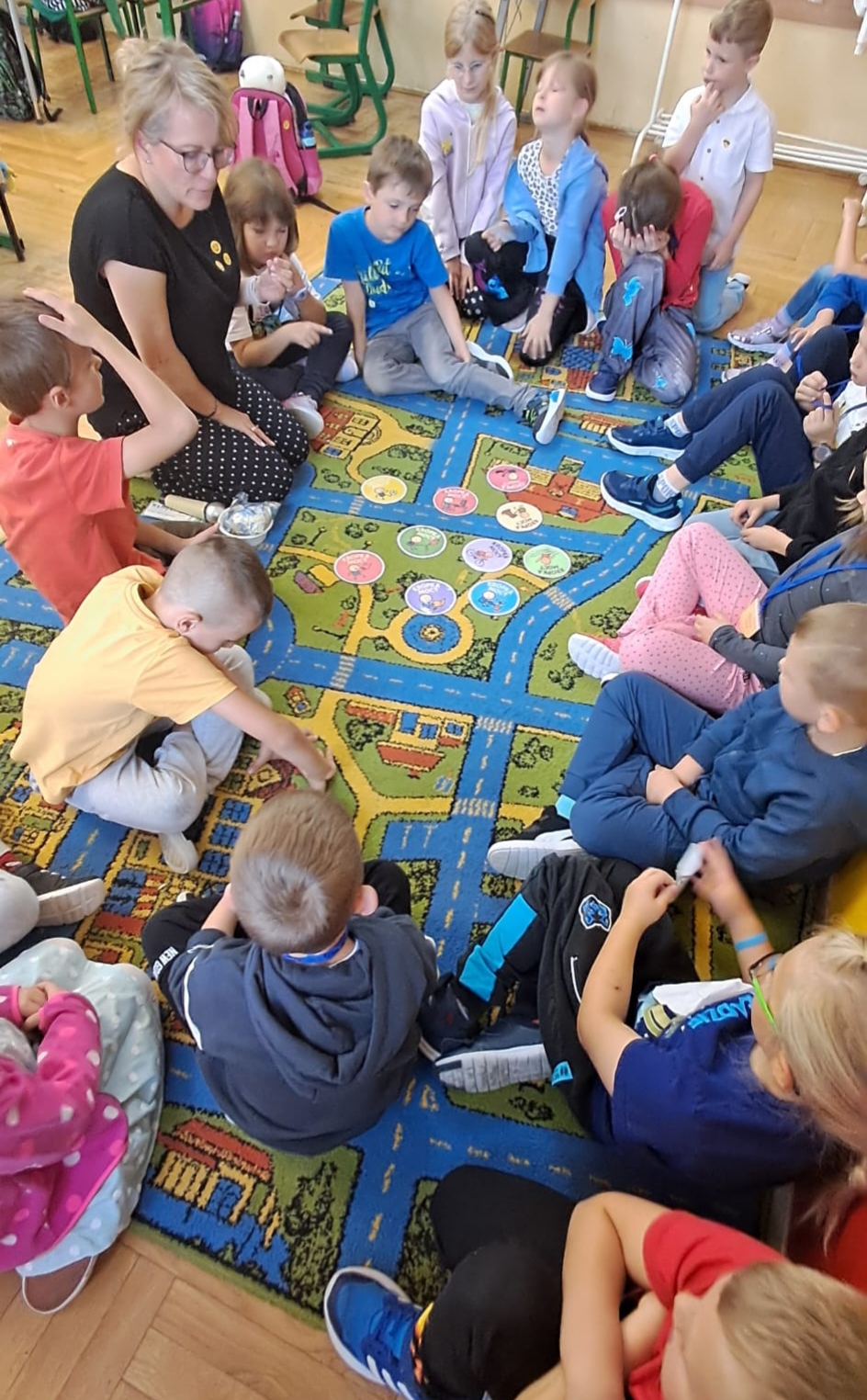  Dzieci siedzą razem z nauczycielką  na dywanie wokół  kolorowych kropek, na krórych narysowane sa różne talenty, np. talent do  gotowania, uprawiania sportów, czytania, malowania itp.