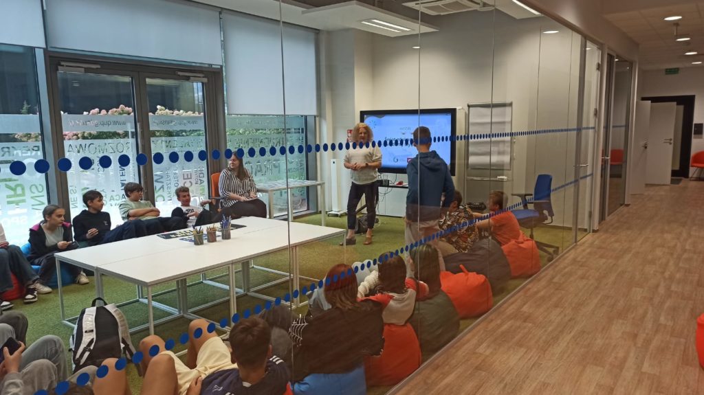 Uczniowie klasy 8c uczestniczą w zajęciach warsztatowych w nowoczesnej sali z przeszklonymi ścianami. Uczestnicy siedzą na kolorowych pufach i krzesłach, a prowadząca stoi obok ekranu mul-timedialnego, na którym wyświetlana jest prezentacja.