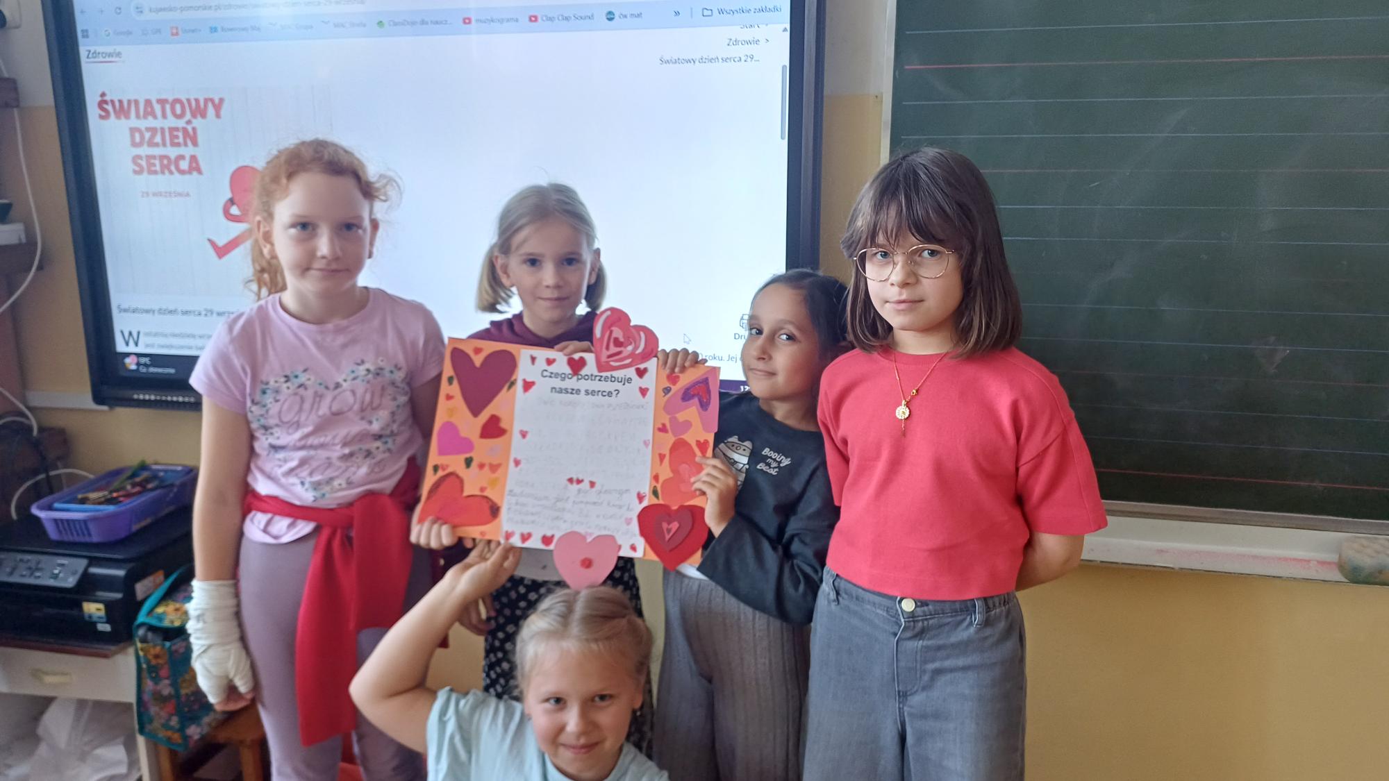 5.DS25 W sali lekcyjnej na tle tablicy multimedialnej stoi 5 uczennic trzymających w dłoni plakat o sercu.