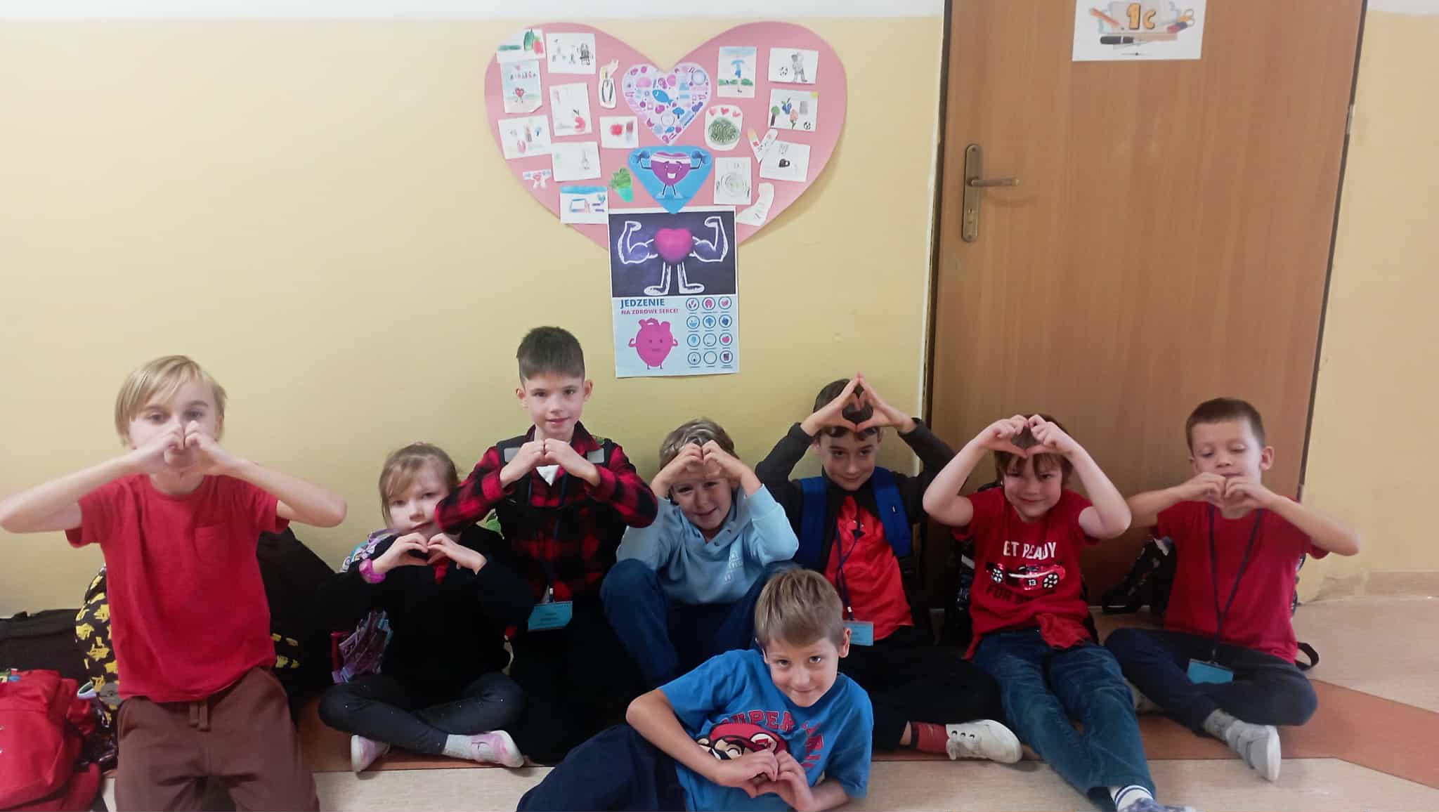 Na tle ściany z plakatem w kształcie serca siedzi ośmioosobowa grupa uczniów klasy 1c z ułożonymi w kształcie serca rączkami.