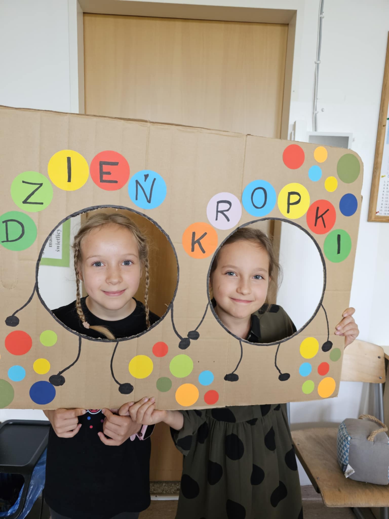 Dwie uśmiechnięte dziewczynki pozują z kolorową dekoracją  z napisem " Dzień Kropki"