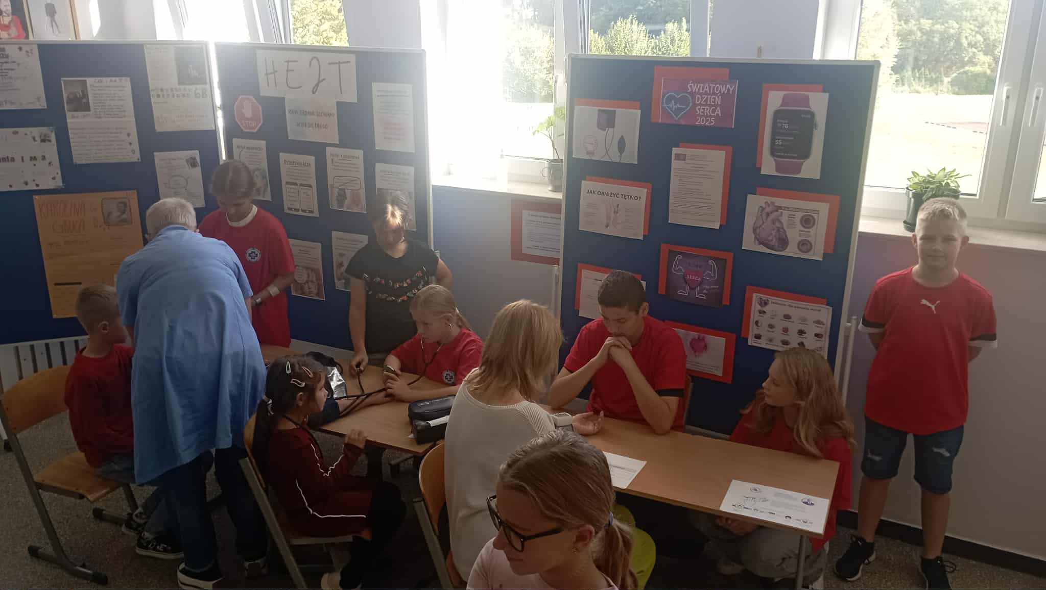 Na półpiętrze szkoły przy stoliku z ciśnieniomierzem na ręku siedzi dyrektor szkoły i 2 uczniów z 1 klasy. Przed stolikiem pielęgniarka szkolna, 6 uczniów Koła PP HOPR. W tle tablica z plakatami o hejcie.