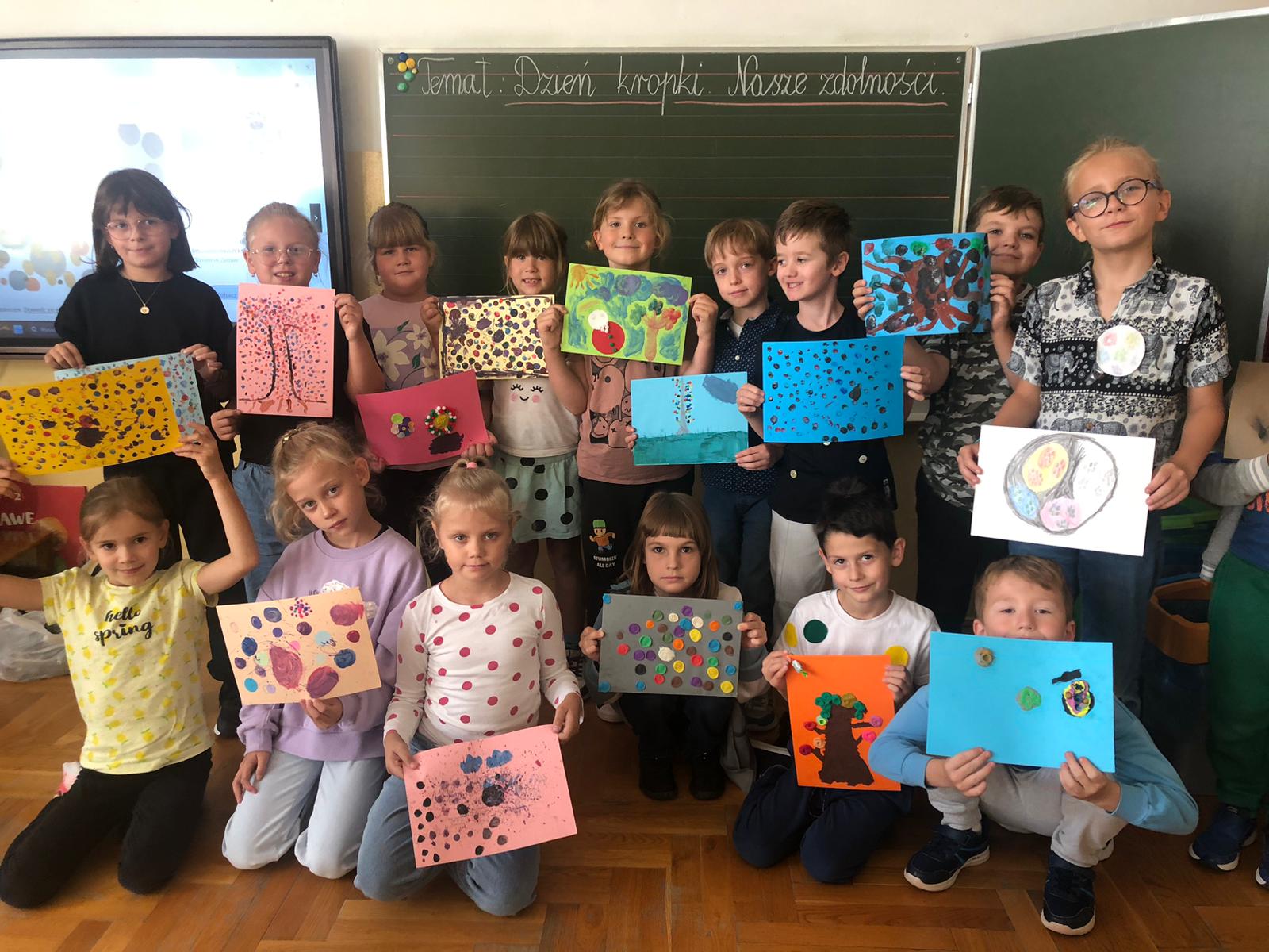 Grupa dzieci w sali lekcyjnej ustawiona jest w dwóch rzędach. Każde z dzieci trzyma w dłoniach pracę plastyczna przedstawijącą  różne kropki. W tle znajduje się duży ekran i tablica.