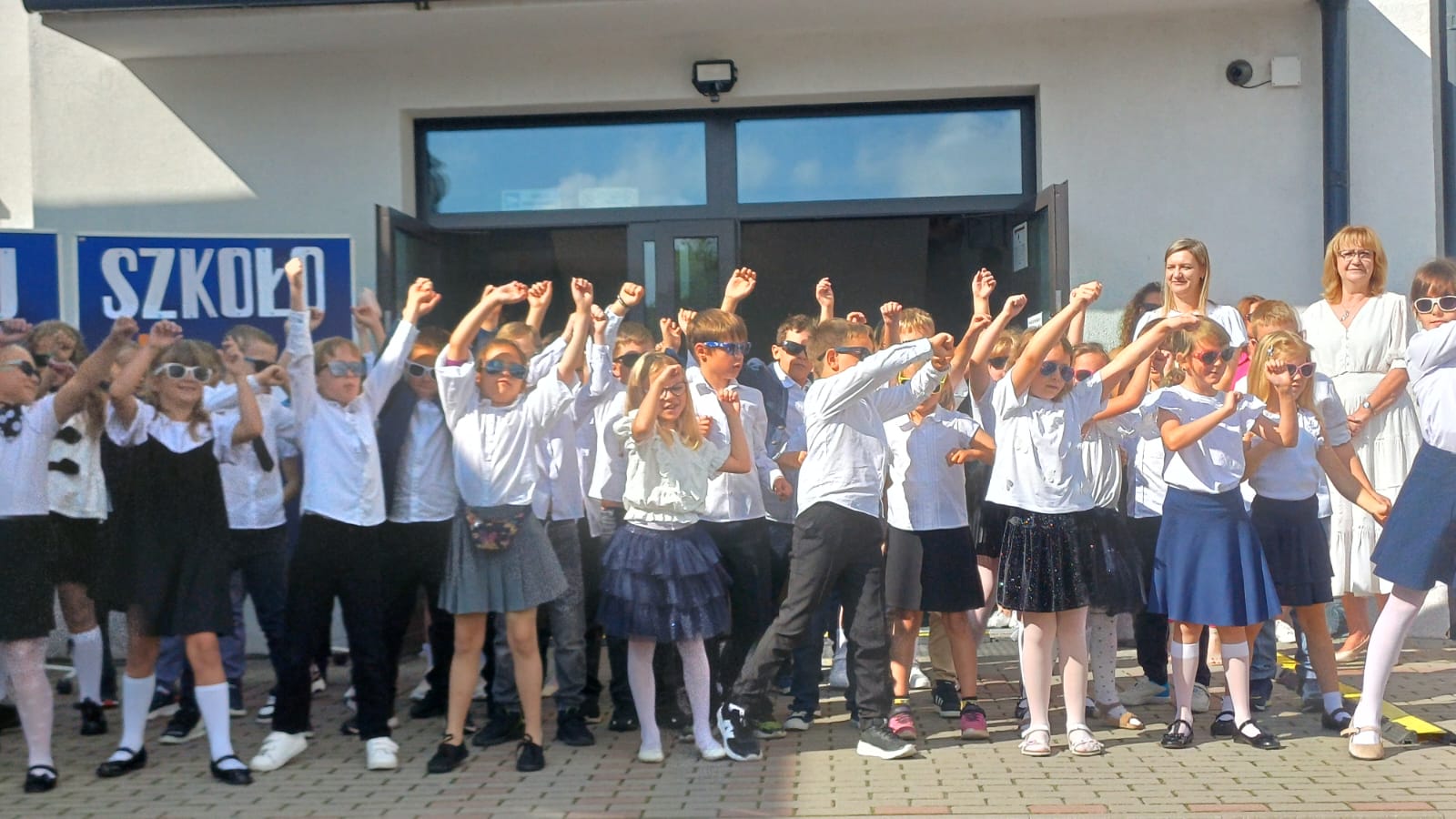 Grupa dzieci ubranych na galowo stoi z podniesionymi rękami z tylu znajdują się nauczyciele.