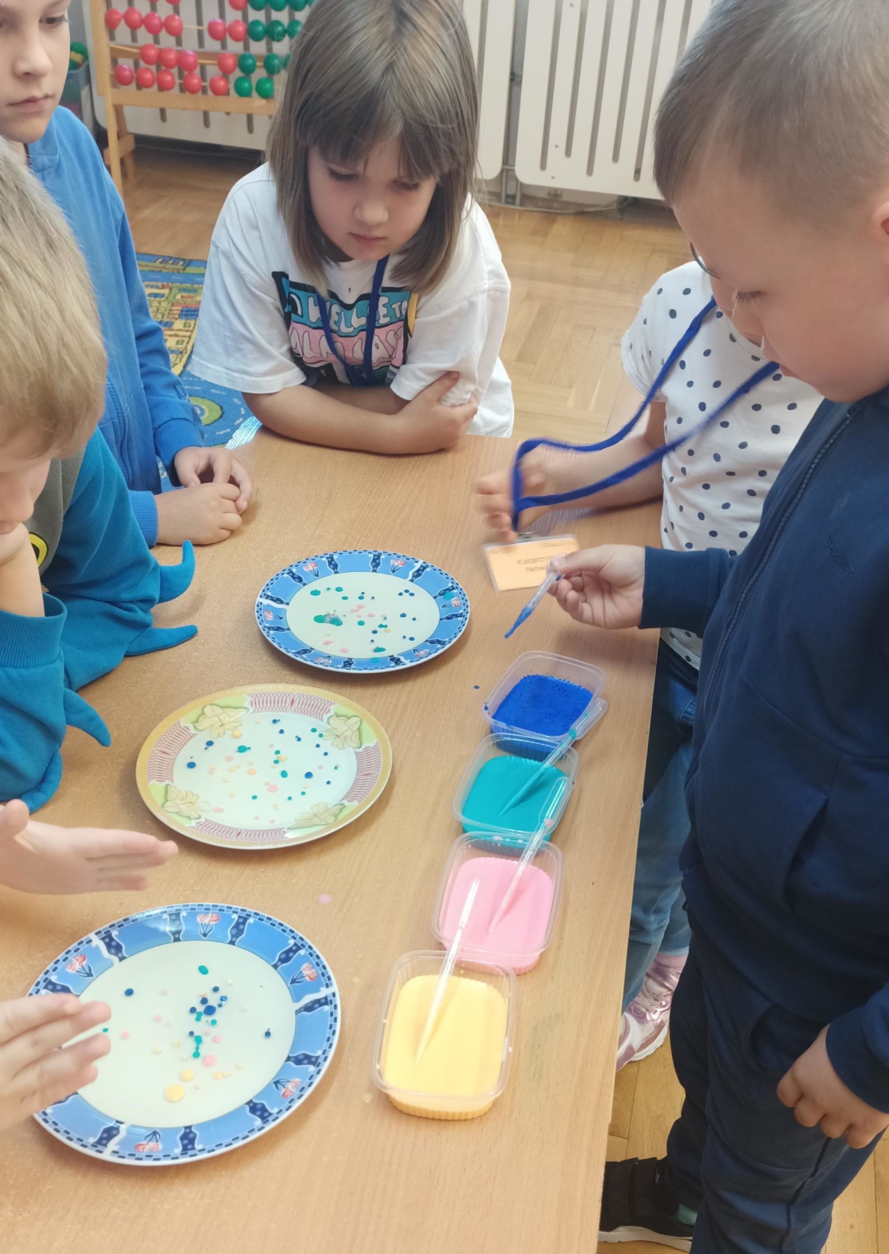   Pięcioro dzieci stoi wokól szkolnej ławki, na której znajdują się trzy talerzyki i trzy pojemniki z kolorową cieczą oraz pipety. Uczniowie obserwują przebieg doświadczenia  z gęstością cieczy.