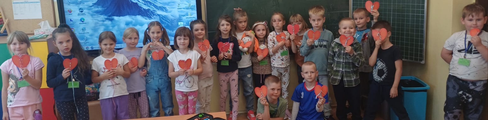 Dzieci z klasy 1b siedzą lub stoją w grupie z serduszkami w rękach. Za dziećmi znajduje się tablica multimedialna.