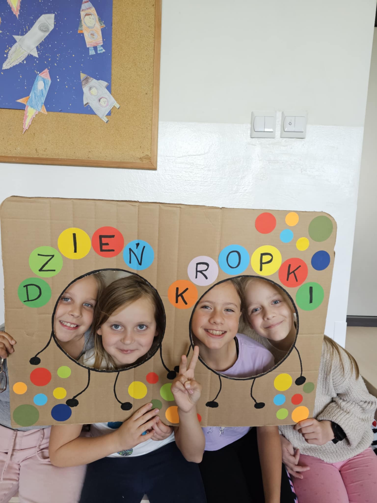 Cztery uśmiechnięte dziewczynki pozują z kolorową dekoracją z napisem " Dzień Kropki". W tle tablica przedstawiająca pojazdy kosmiczne.