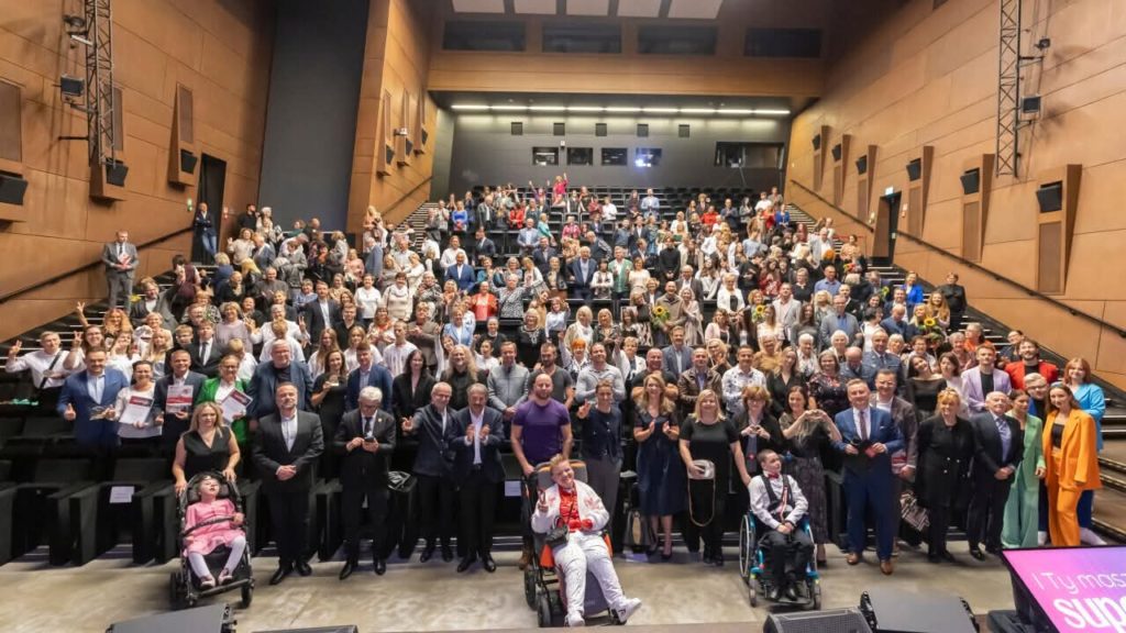 Duża grupa osób pozuje wspólnie do pamiątkowego zdjęcia na scenie i widowni sali audytoryjnej. Wśród uczestników są osoby w różnym wieku, także osoby z niepełnosprawnościami, w tym na wózkach. Atmosfera jest radosna i uroczysta.