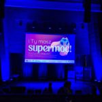 Oświetlona scena z dużym ekranem projekcyjnym, na którym widnieje napis: „I Ty masz supermoc! Bursztynowe Dźwigi 2025”. Widzowie powoli zajmują miejsca przed rozpoczęciem wydarzenia.