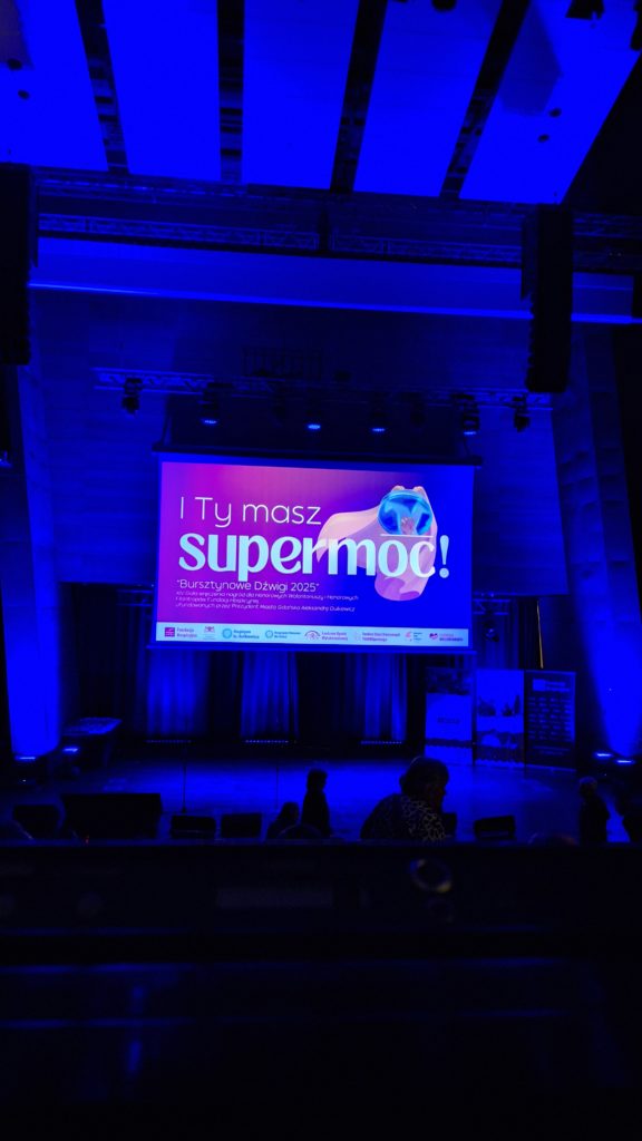 Oświetlona scena z dużym ekranem projekcyjnym, na którym widnieje napis: „I Ty masz supermoc! Bursztynowe Dźwigi 2025”. Widzowie powoli zajmują miejsca przed rozpoczęciem wydarzenia.