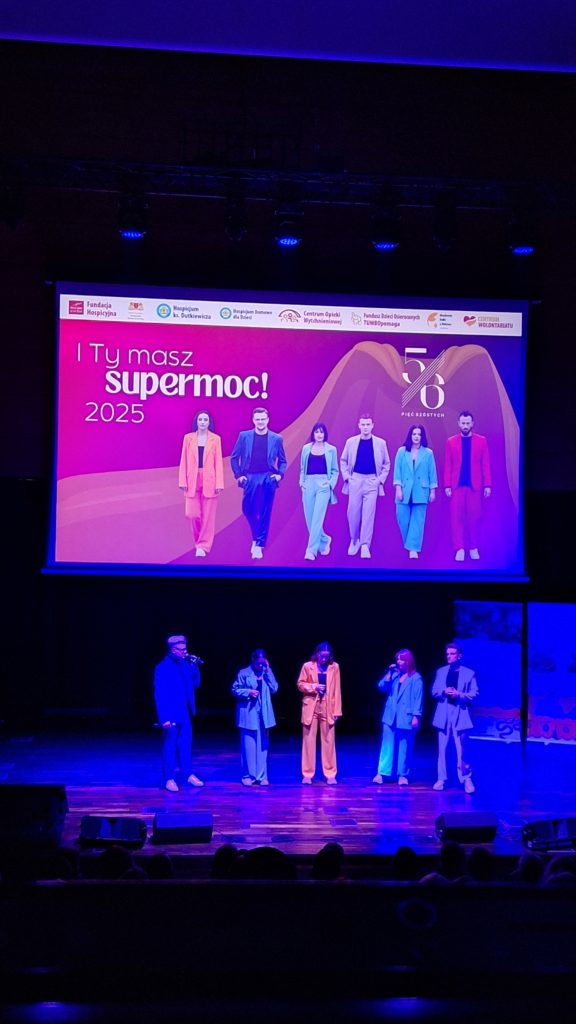 Na scenie stoi pięć osób w kolorowych garniturach, śpiewających do mikrofonów. Nad nimi wisi duży ekran z grafiką kampanii „I Ty masz supermoc! 2025”.