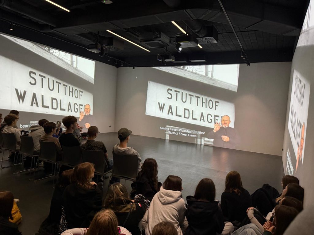 W sali na trzech ścianach wyświetlany jest film dotyczący historii obozu Stutthof. Na środku sali oraz pod jedną ściana na podłodze siedzą uczniowie oglądając przedstawiany materiał.