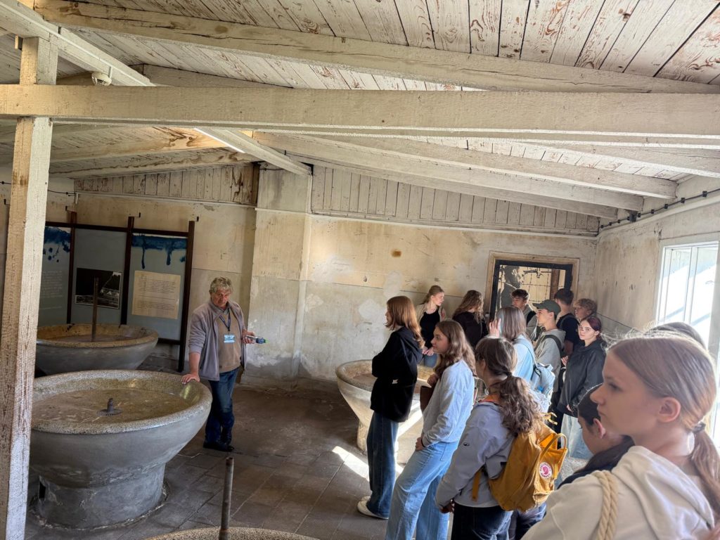 W pomieszczeniu będącym łaźnią więźniarską uczniowie stoją przy okrągłych betonowych misach. Na środku stoi przewodnik opierając się o jedną z mis.
