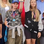 Dwie dziewczyny stoją obok siebie i trzymają w rękach duże puchary. Uśmiechają się, wyglądają na zadowolone i dumne. Ubrane są w sportowe stroje.