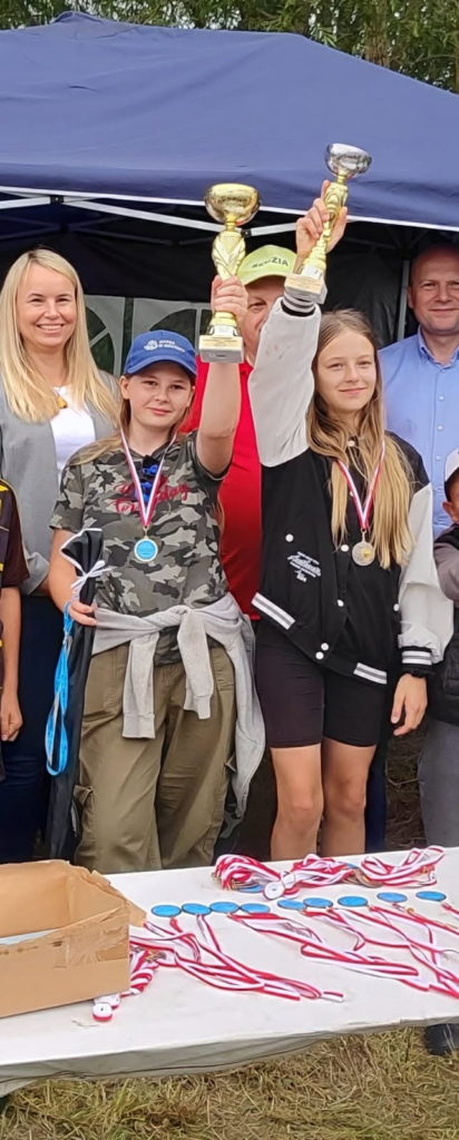 Dwie dziewczyny stoją obok siebie i trzymają w rękach duże puchary. Uśmiechają się, wyglądają na zadowolone i dumne. Ubrane są w sportowe stroje.