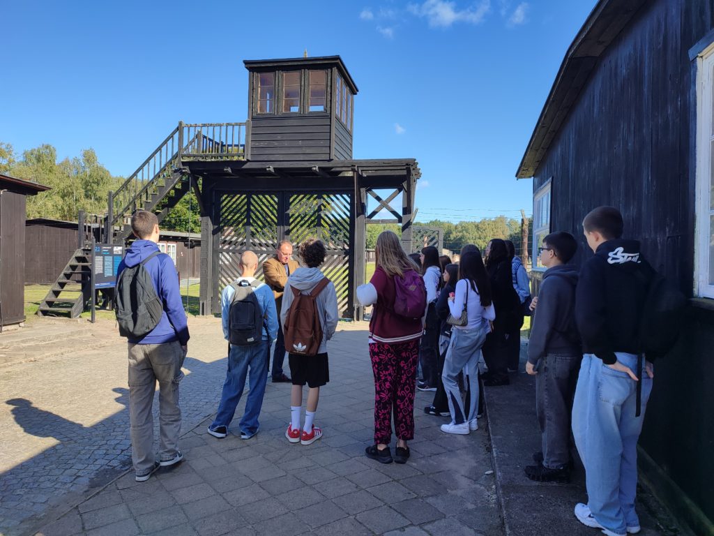 W słoneczny dzień grupa młodzieży stoi przed bramą oddzielającą dwie części muzeum. Brama jest drewniana, wysoka, po bokach otoczona siatką z kolczatką.