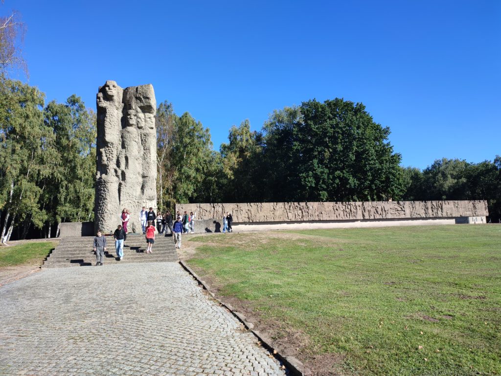 o lewej stronie jest prosta ścieżka prowadząca do ogromnego, kamiennego pomnika w tle. Po prawej stronie, przed pomnikiem jest duży trawnik. Pomnik z tyłu jest jasny, po lewej stronie zdjęcia pionowy, tuż po nim stoją zapalone znicze. Całość pomnika ciągnie się za pionowym elementem od lewej do prawej strony, poziomy wielki posąg wyryte ma daty oraz narodowości osób poległych.