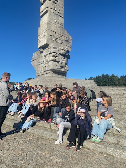 Grupa młodych osób siedzących na schodach pomnika. W tle znajduje się monumentalna budowla z kamienia — to Pomnik Obrońców Wybrzeża na Westerplatte w Gdańsku.
