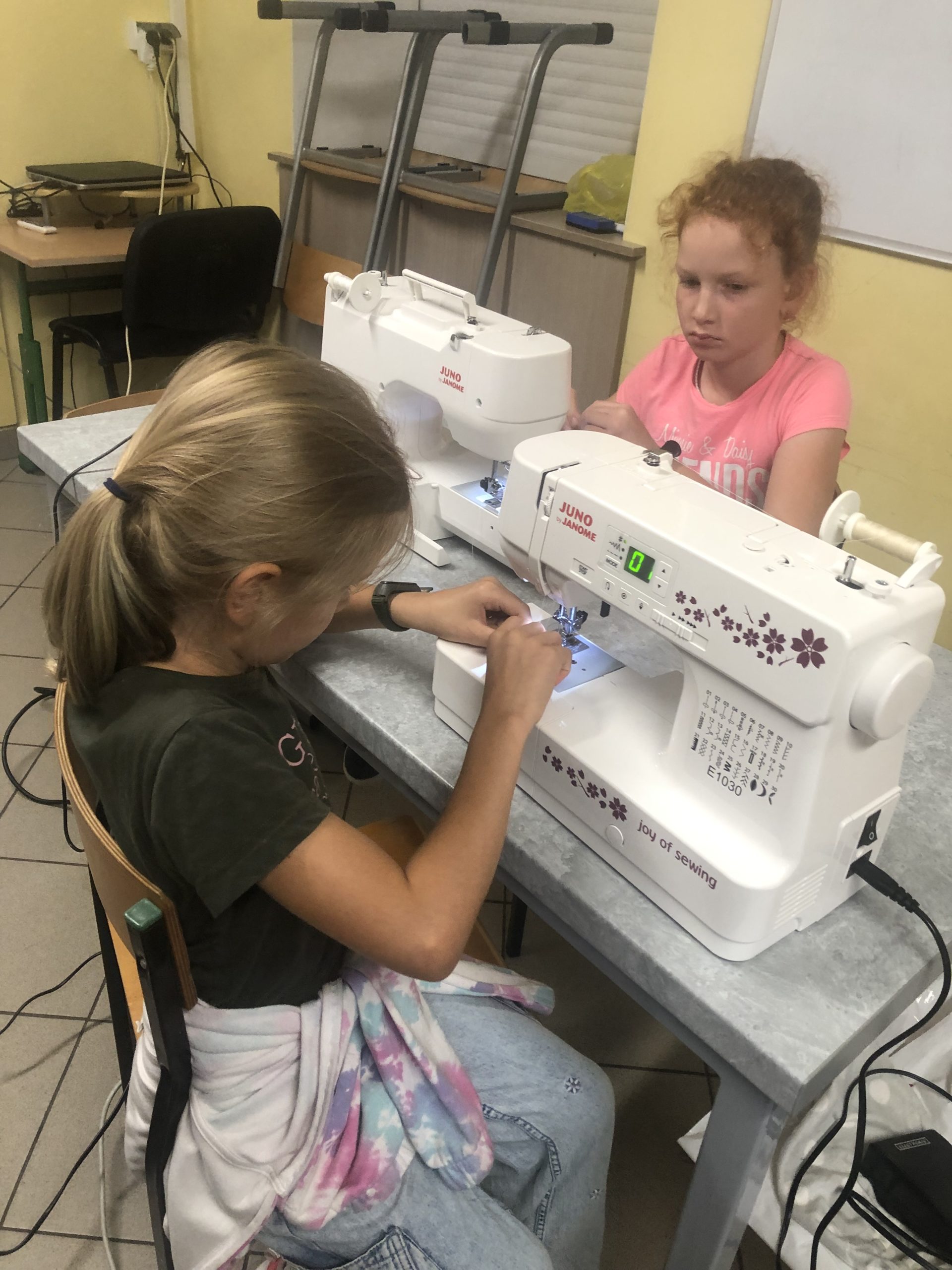 Dwie dziewczynki siedzą przy stole, przed nimi znajdują się maszyny do szycia.