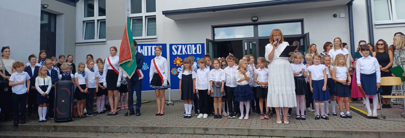 Grupa dzieci i nauczycieli stoi na schodach wejścia do szkoły, przed nimi stoi Pani Dyrektor z mikrofonem.