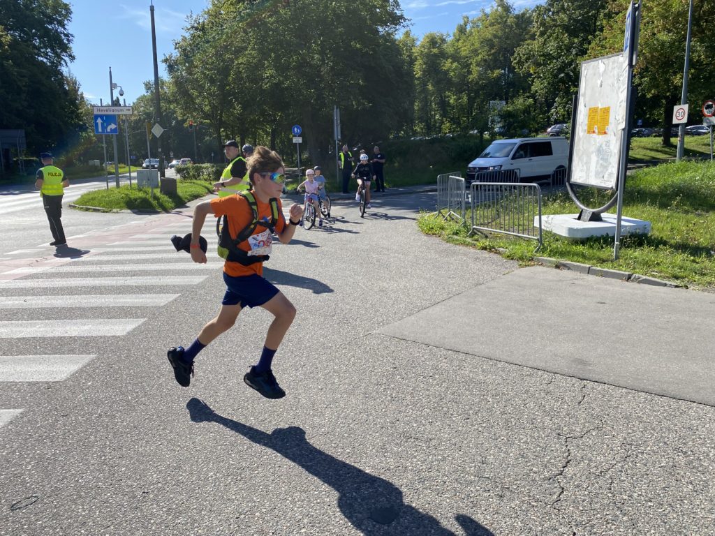 Chłopiec w stroju sportowym i okularach sportowych biegnie ulicą podczas maratonu. Przy trasie stoją policjanci zabezpieczający trasę biegu.