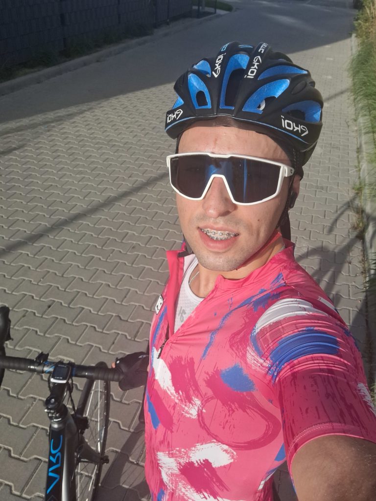 Mężczyzna w kasku rowerowym i okularach sportowych robi selfie. Stoi przy rowerze i ma na sobie koszulkę kolarską.
