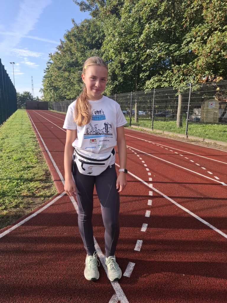 Uśmiechnięta dziewczynka w stroju sportowym stoi na bieżni. W tle są drzewa i świeci słońce.