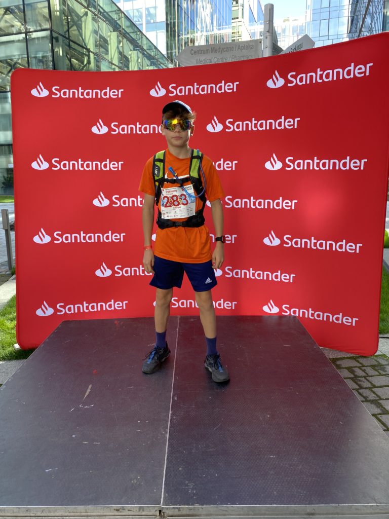 Chłopiec w stroju sportowym i okularach sportowych stoi na tle baneru Santander Bank, organizatora biegu charytatywnego. Na koszulce ma nr startowy 283.