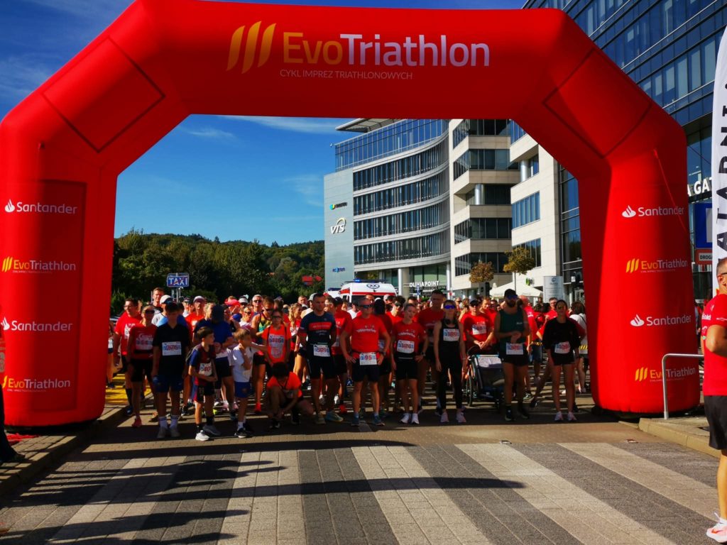 Uczestnicy biegu stojący na starcie pod dużą bramą z napisem „EvoTriathlon”. W tle nowoczesne budynki i słoneczna pogoda.