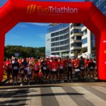 Uczestnicy biegu stojący na starcie pod dużą bramą z napisem „EvoTriathlon”. W tle nowoczesne budynki i słoneczna pogoda.
