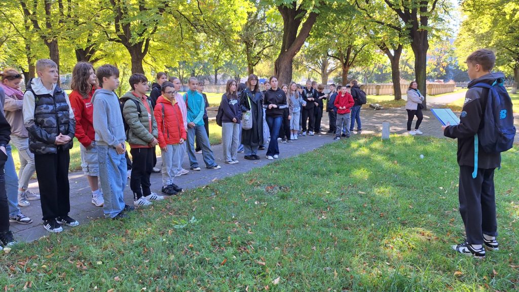 Grupa uczniów stoi w parku, uważnie słuchając chłopca trzymającego kartkę, z której czyta. Wokół nich widać drzewa
