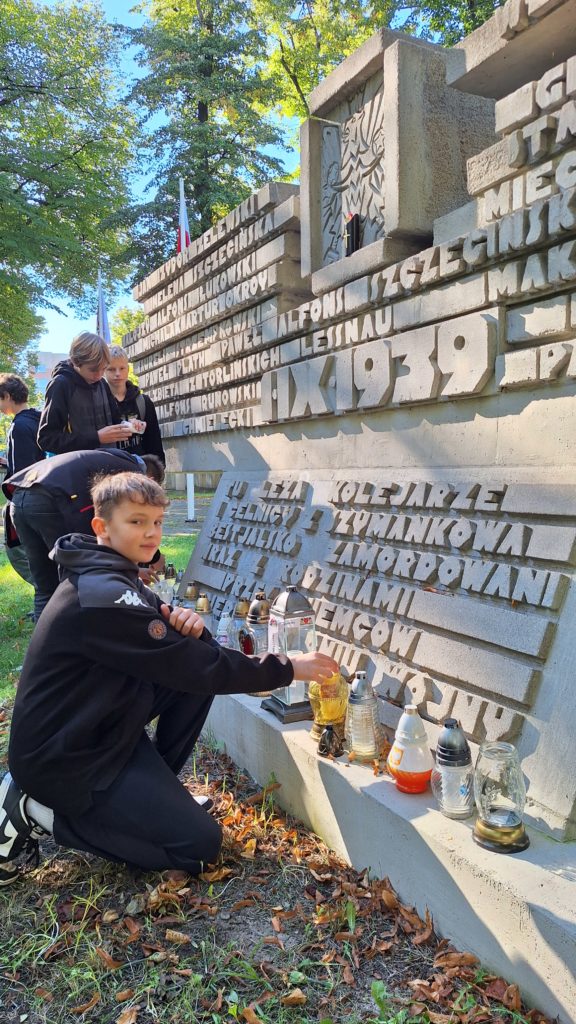 Chłopiec zapala znicz przed betonowym pomnikiem upamiętniającym ofiary II wojny światowej, obok niego stoją inni uczniowie.