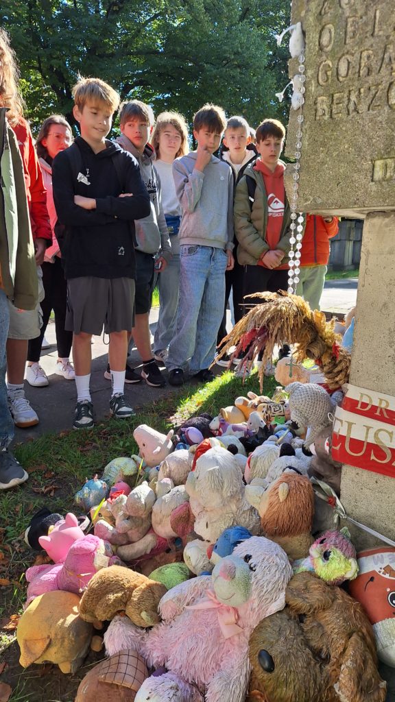 Grupa uczniów stoi przed pomnikiem, który jest przykryty licznymi pluszowymi zabawkami, takimi jak misie i inne pluszaki. Dzieci cicho przyglądają się wystawie, niektóre patrzą na pomnik.