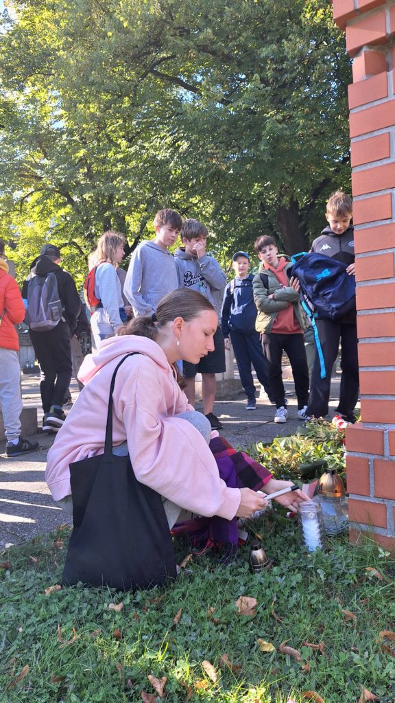 Dziewczyna zapala znicz przy grobie, w tle grupa uczniów obserwuje.