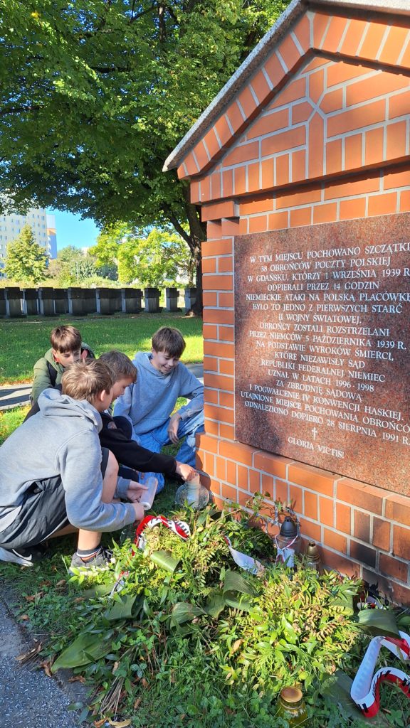 Grupa chłopców klęczy wokół pomnika, zapalając znicze i składając kwiaty u jego podstawy. Pomnik upamiętnia polskich pocztowców, którzy walczyli podczas niemieckiej inwazji w 1939 roku.