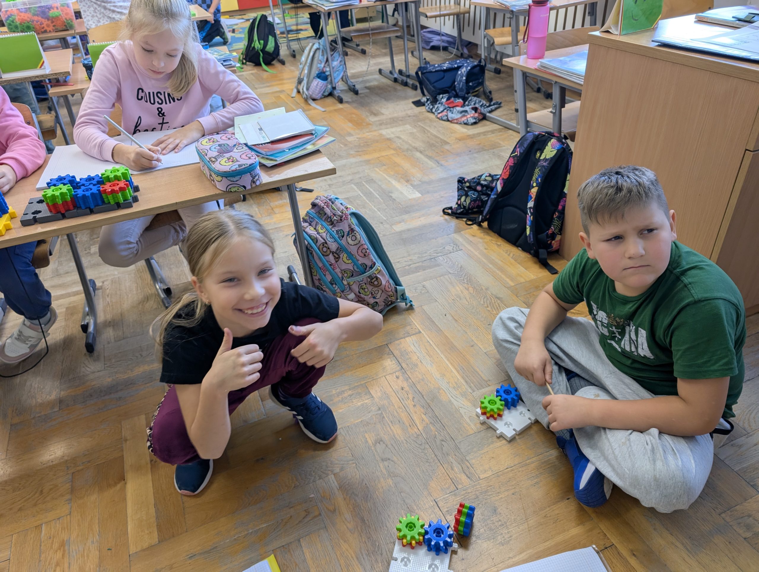 Uczniowie w klasie podczas zajęć z programu „Kolorowa Matematyka z Korbo”. Dziewczynka pokazuje kciuki w górę, obok niej chłopiec układa kolorowe klocki zębate Korbo.