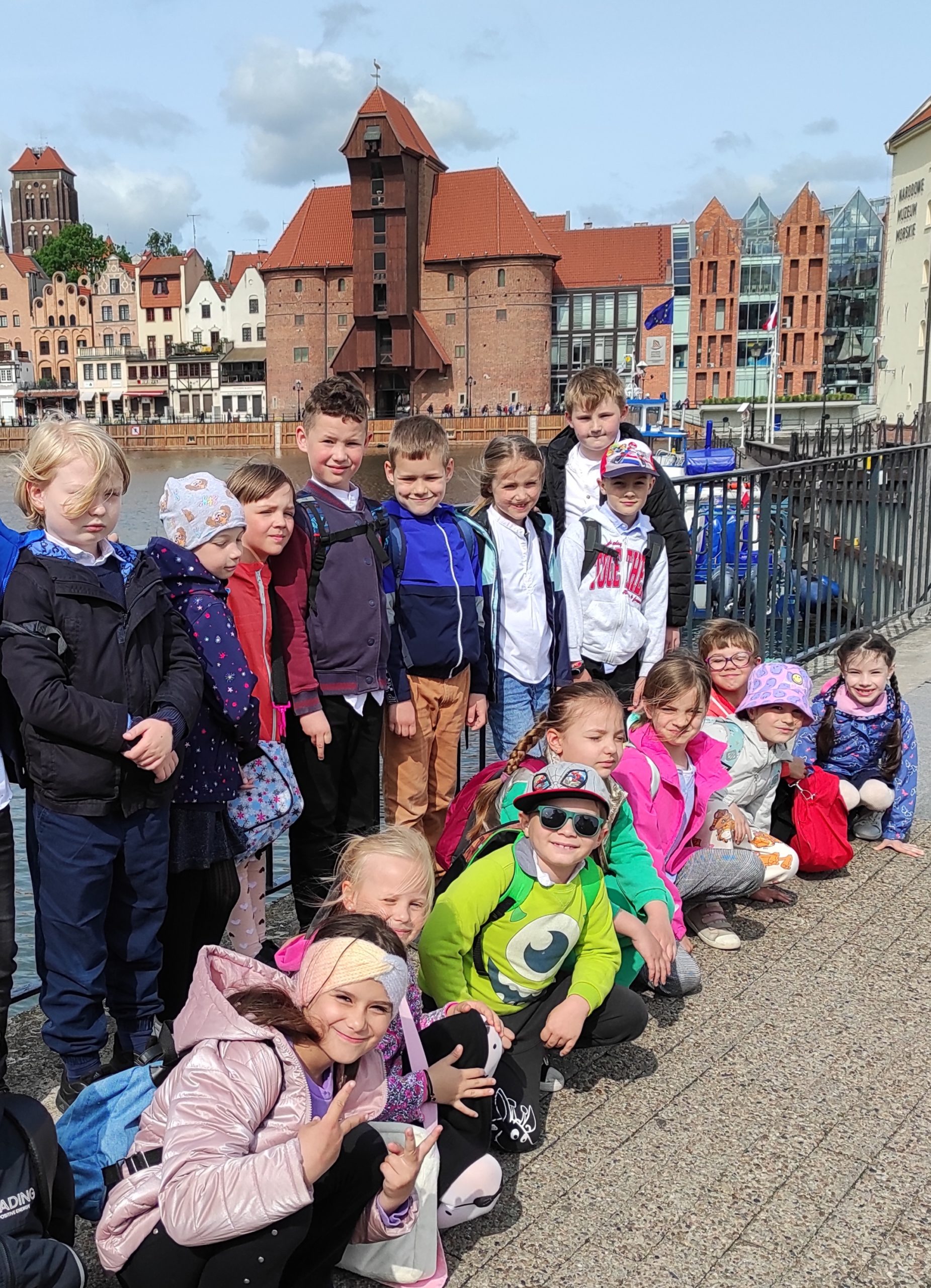 Zdjęcie wykonane w plenerze. Na pierwszym planie dzieci ustawione w dwa rzędy. Jeden stoi, drugi kuca. W tle Żuraw oraz inne budynki.