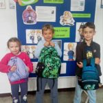 Na szkolnym korytarzu na tle tablicy z napisem Ogólnopolski Dzień Tornistra stoi uczennica i dwaj uczniowie z plecakami w dłoniach.