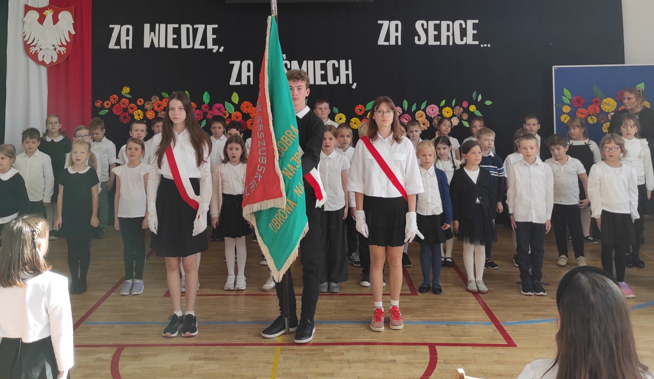 Na pierwszym planie troje nastolatków: chłopiec trzyma sztandar, z jego prawej i lewej strony stoją dwie dziewczyny. Za nastolatkami stoją dzieci w strojach galowych, ustawione w trzech rzędach. W tle dekoracja: zawieszona pionowo flaga Polski, na niej godło,  dalej napis i kwiaty.