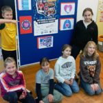 W sali lekcyjnej w koło tablicy z plakatem „Weź udział w rekordzie w pierwszej pomocy” stoją lub kucają uczniowie Koła PP HOPR.