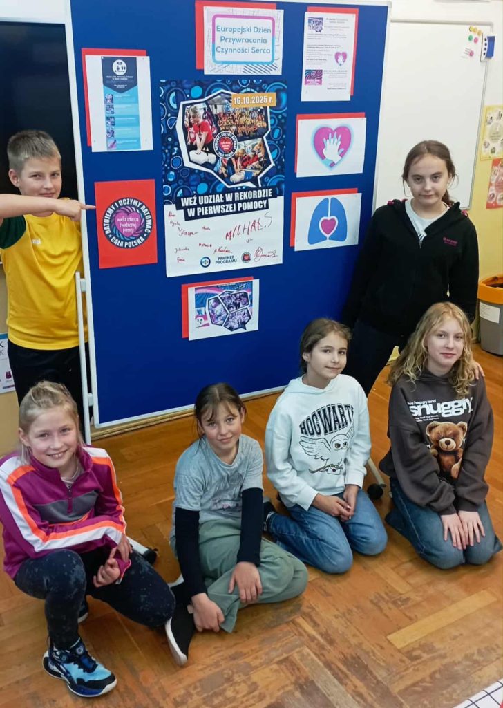 W sali lekcyjnej w koło tablicy z plakatem „Weź udział w rekordzie w pierwszej pomocy” stoją lub kucają uczniowie Koła PP HOPR.