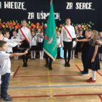 W sali gimnastycznej na tle dekoracji z kwiatami stoi 6 uczniów klas pierwszych z wyciągniętymi w stronę sztandaru palcami do ślubowania. W tle stoi poczet sztandarowy oraz pozostali uczniowie klas pierwszych.