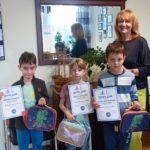 W gabinecie Dyrektora stoi jedna dziewczynka i dwóch chłopców z nagrodami i dyplomami w rękach. Przy nich stoi Pani Dyrektor. W tle krzesła, lustro oraz komoda, na której leży kwiat w doniczce i drewniany statek.