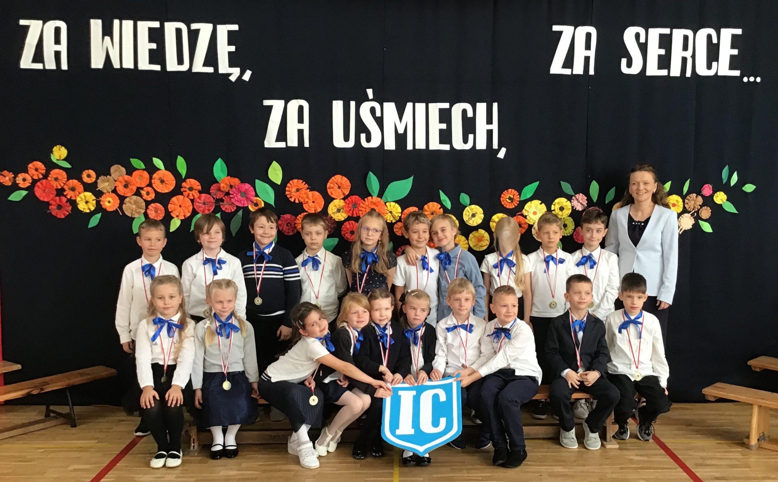Na tle kwiatowej dekoracji i napisu stoją lub siedzą na ławeczce uczniowie klasy IC wraz z wychowawcą.