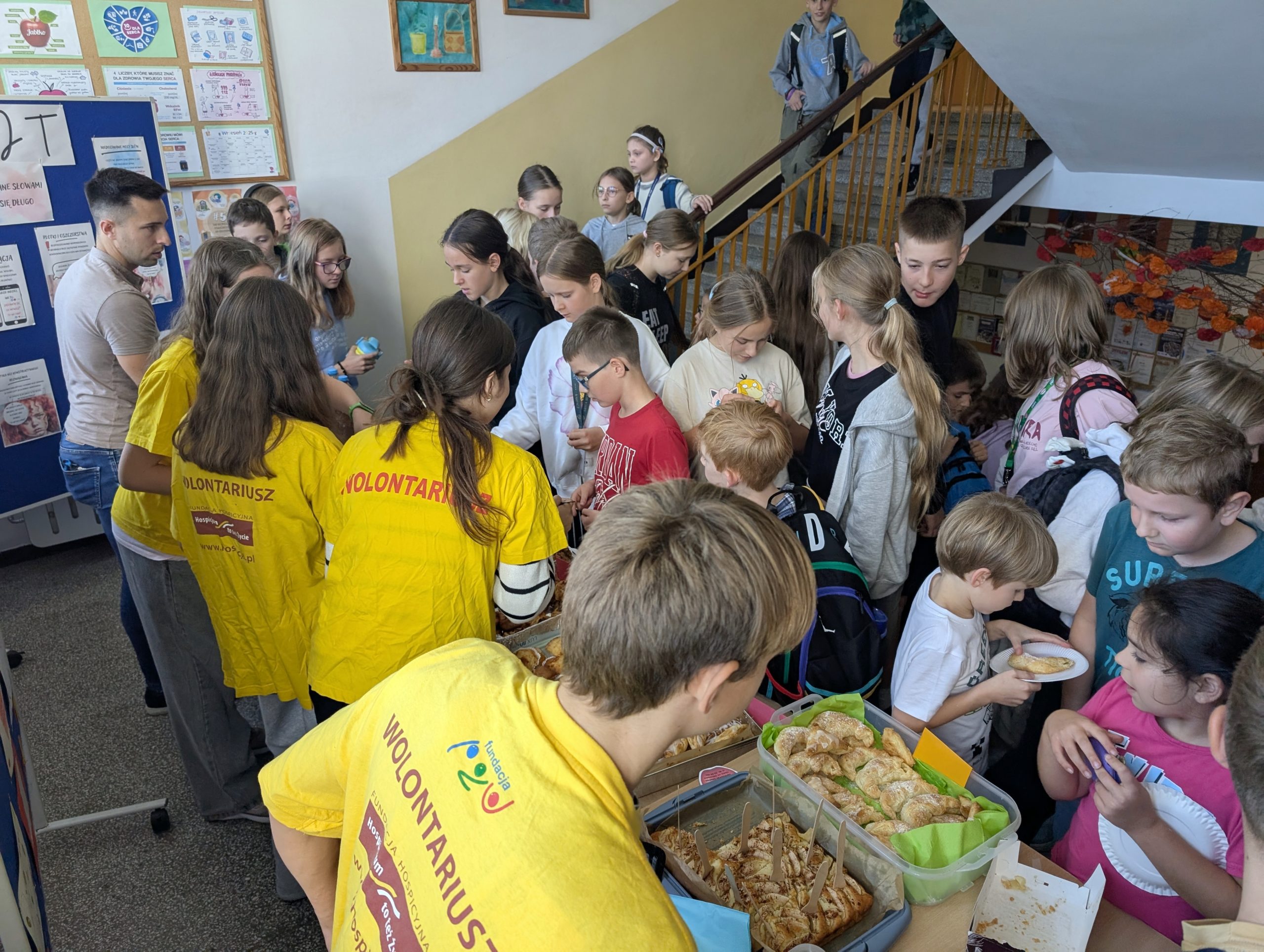 Stoisko z wystawionymi ciastami z jabłek, obsługiwane przez 4wolontariuszy, przed którymi stoi grupa dzieci. za wolontariuszami stoi ich koordynator.