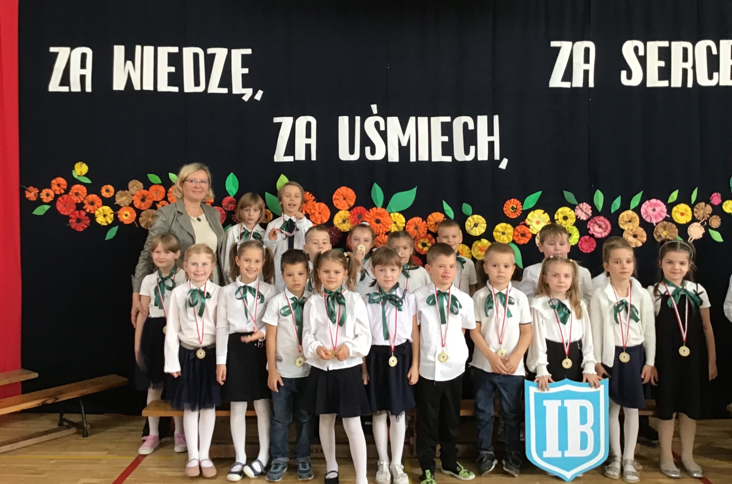 Na tle kwiatowej dekoracji i napisu stoją lub siedzą na ławeczce uczniowie klasy IB wraz z wychowawcą.