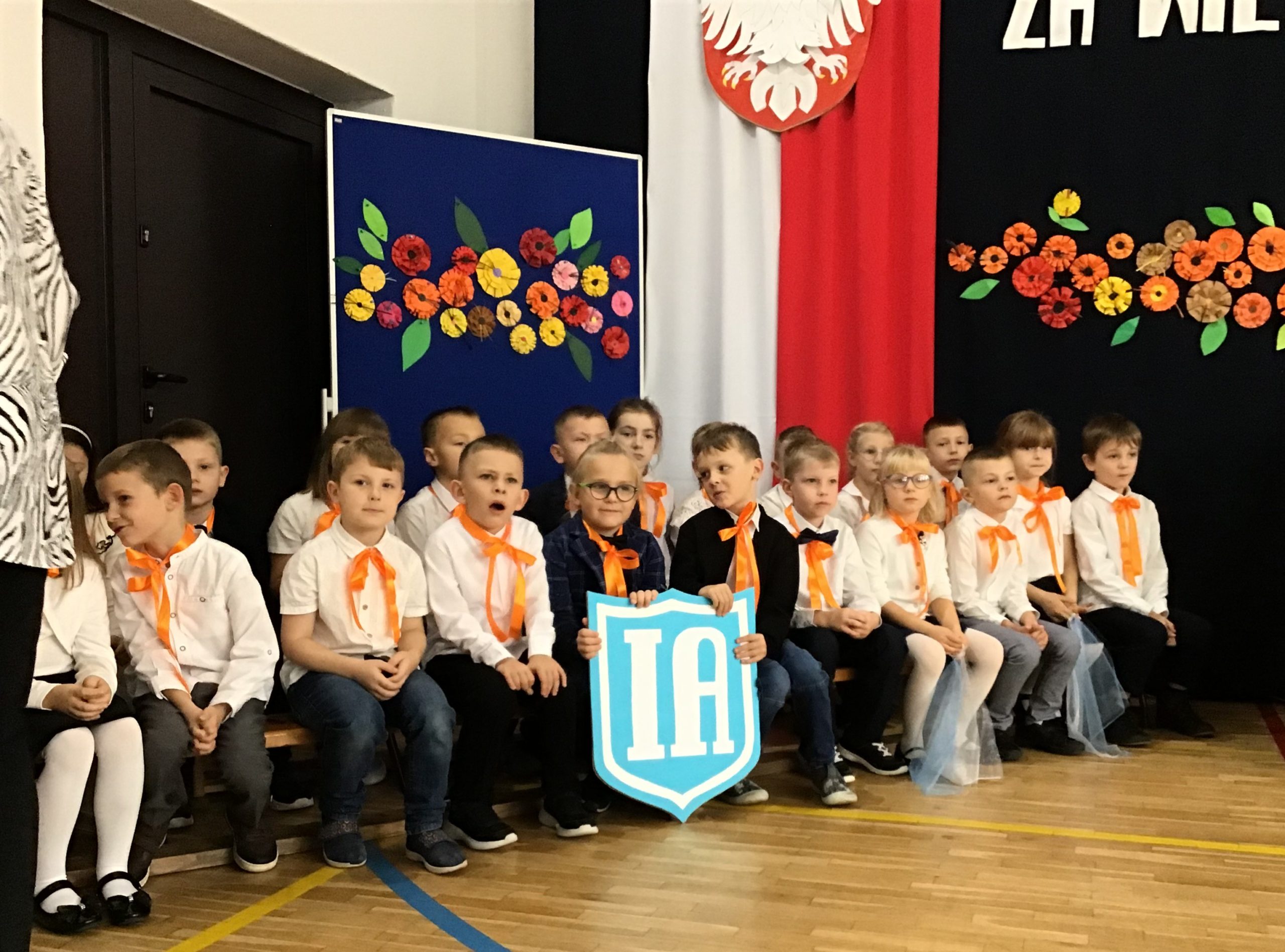 Na tle kwiatowej dekoracji siedzą na ławeczkach uczniowie klasy IA.